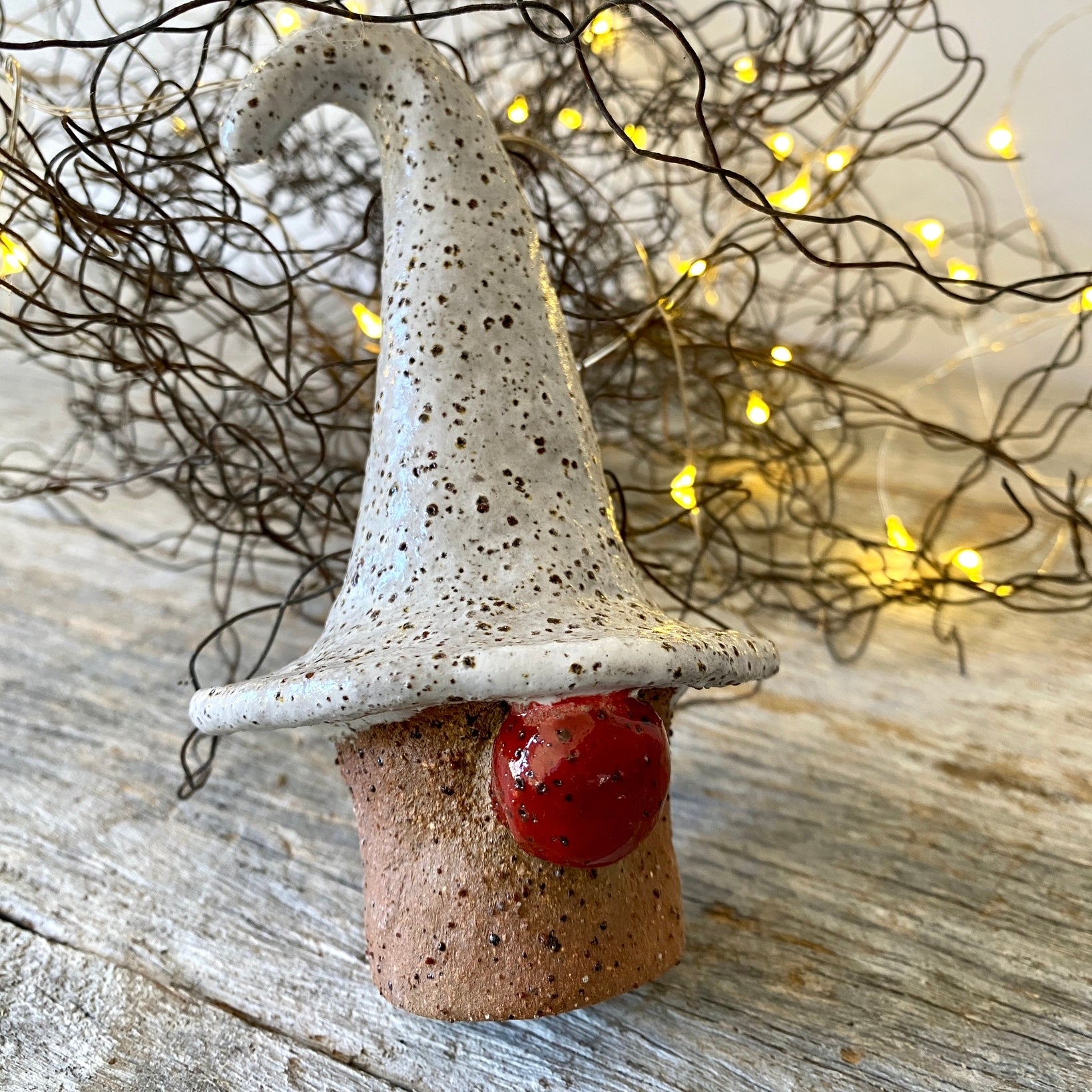 A ceramic Christmas gnome with a white hat and red nose placed on a wooden surface with twinkle lights in the background.