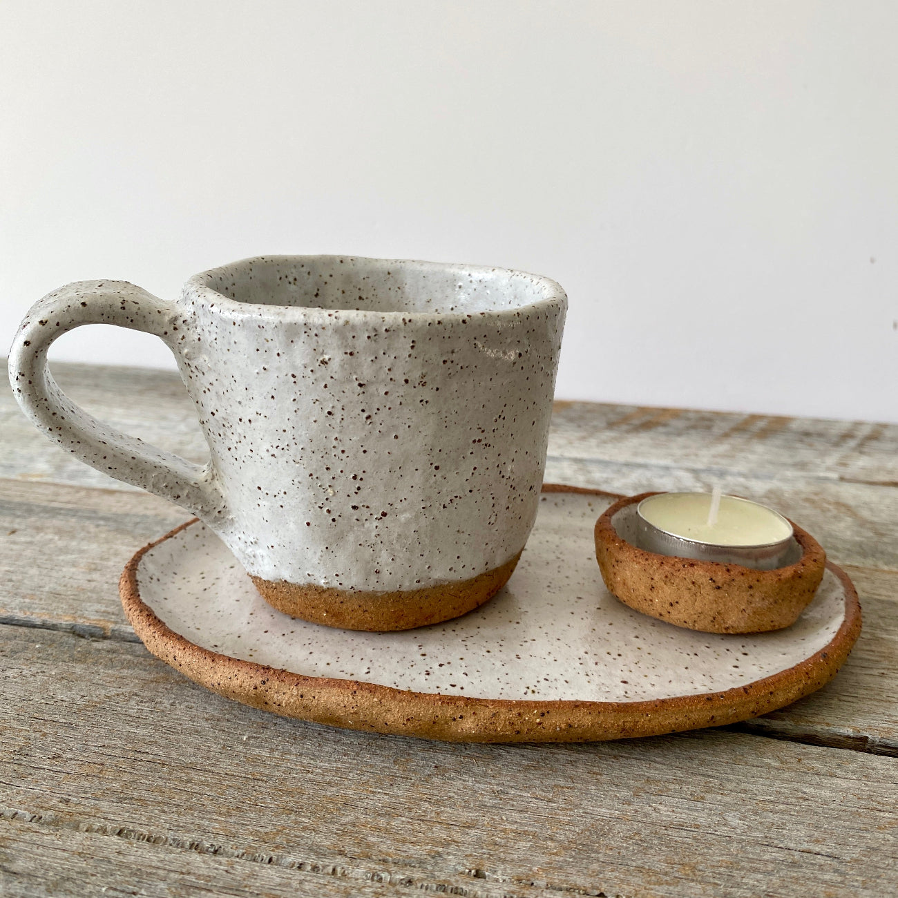 A rustic ceramic tea/coffee cup with a speckled design on a white oval plate, with a small tealight candle holder to the side, all placed on a wooden surface.